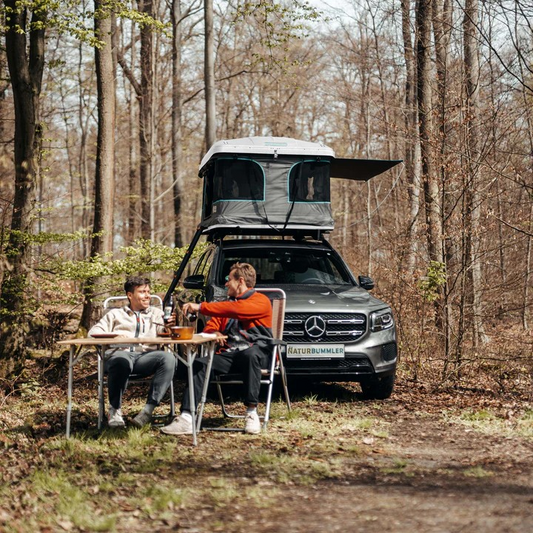 Naturbummler Dachzelt Flitzer 1 Xl mit Hartschale in Weiß bis 4 Prsonen. Zwei Leute chillen entspannt vor ihrem aufgebauten Dachzelt, sitzen am Tisch und lassen sich von der Ruhe des Waldes umhüllen. Kein Stress, kein Lärm-einfach nur der Moment, das Zelt, der Wald und gute Gesellschaft.